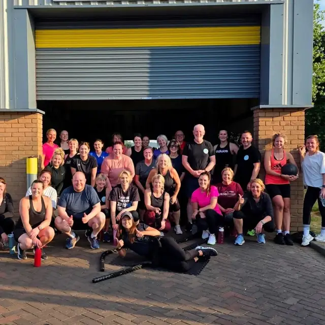 A big, smiling bootcamp crew gathered in front of the raised shutter door of the gym