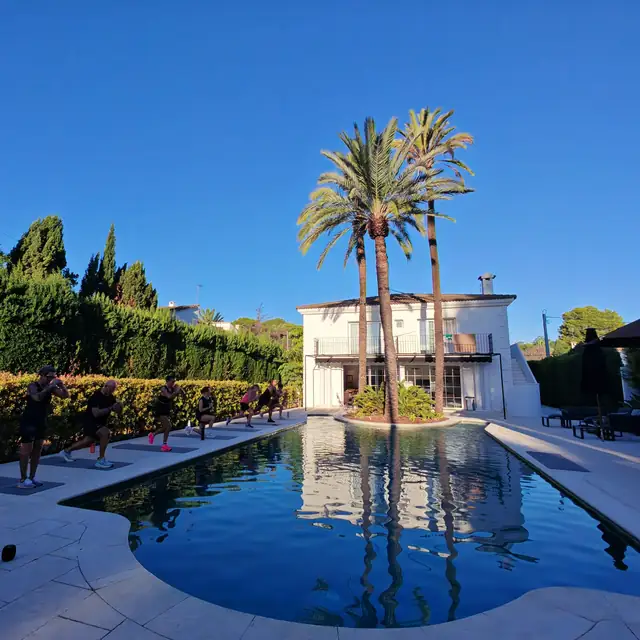 A group working out beside a villa pool under palm trees and blue sky.