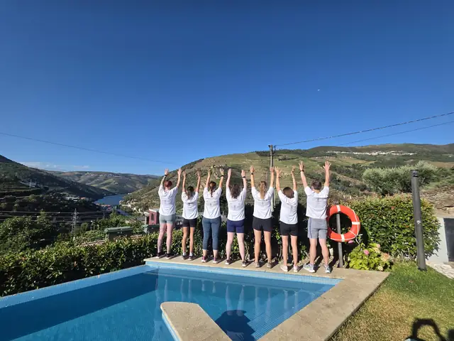 A Let Luce group in white shirts behind a villa pool, arms raised, with terraced hills and the river behind.