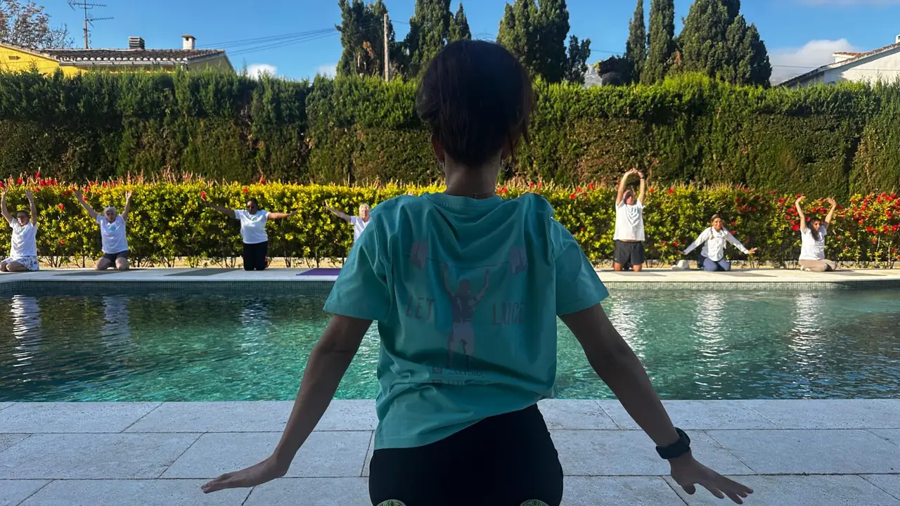 Lucy, seen from behind in her Let Luce shirt, at the edge of the pool, watching the retreat group stretch on mats across the water.