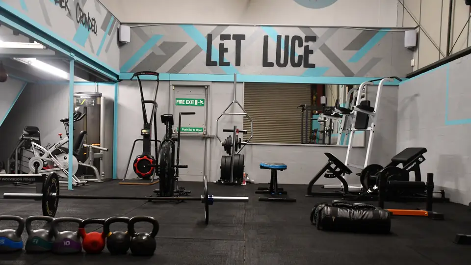Wide view of the training floor at Unit 49 under the LET LUCE sign, with a row of coloured kettlebells, a loaded barbell, a squat rack and rubber flooring.