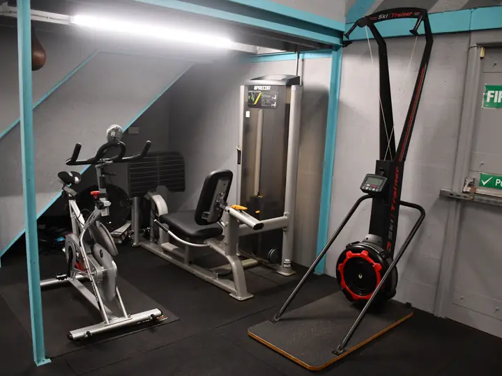 Two exercise bikes, a leg press machine and a red air bike in the corner under a teal steel frame, next to a fire exit door.