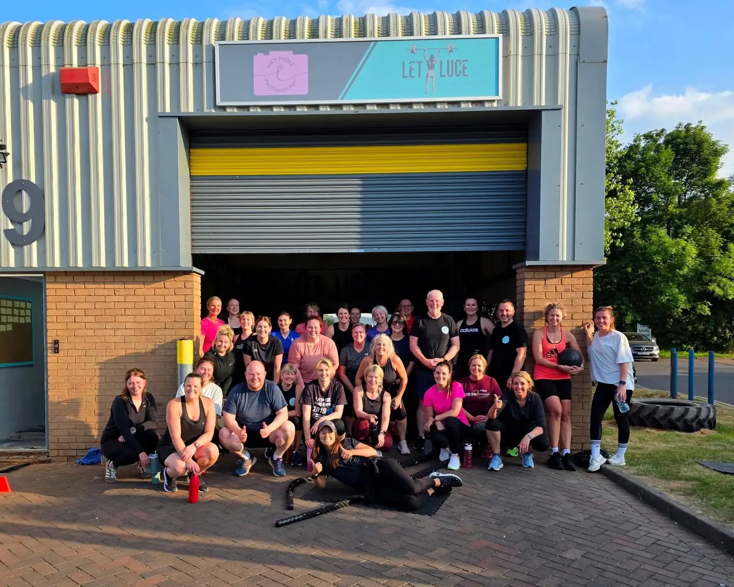 A big, smiling bootcamp crew gathered in front of the raised shutter door of the gym